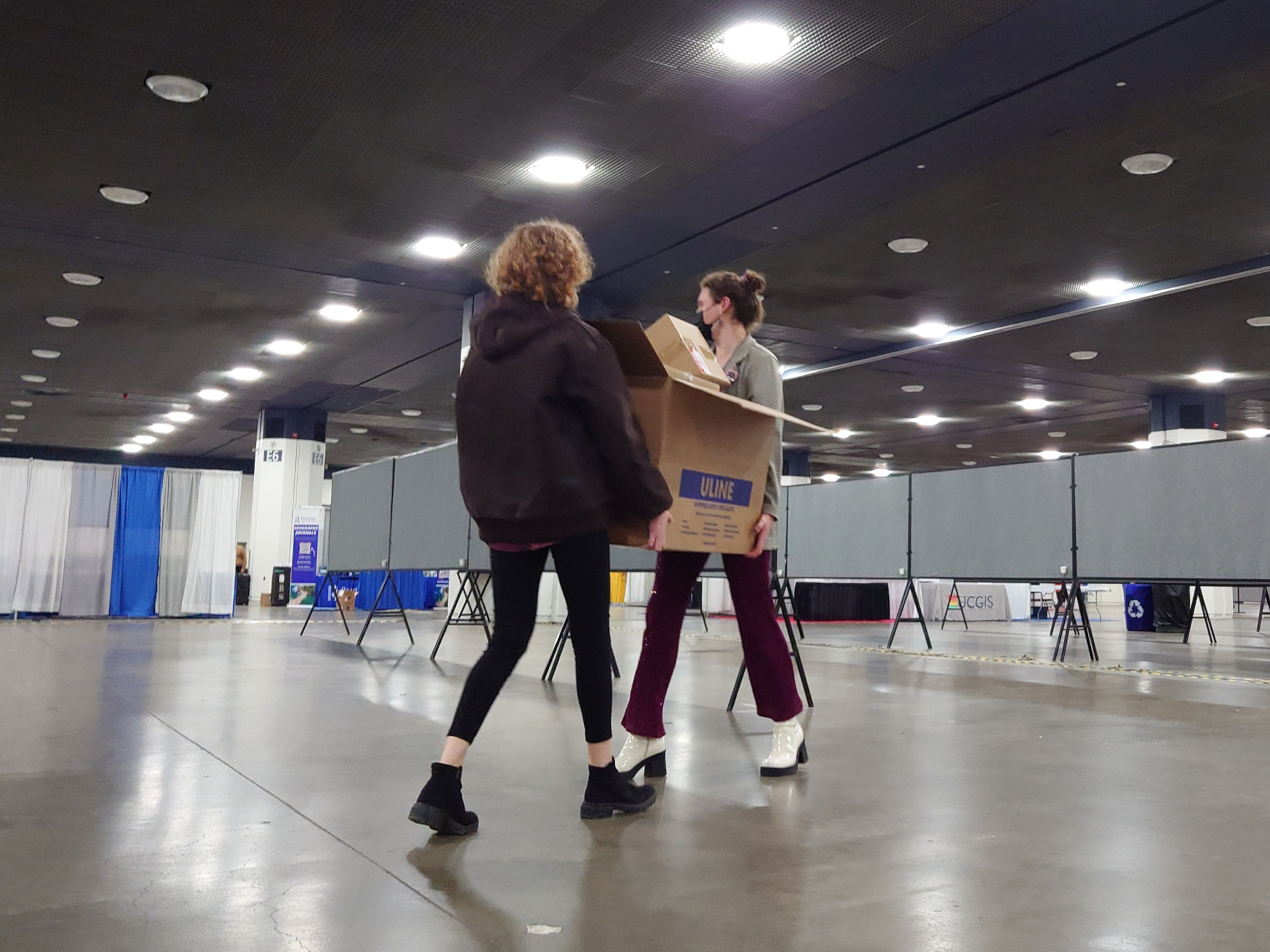 Two mischief makers carry a large box of zines across a conference hall.