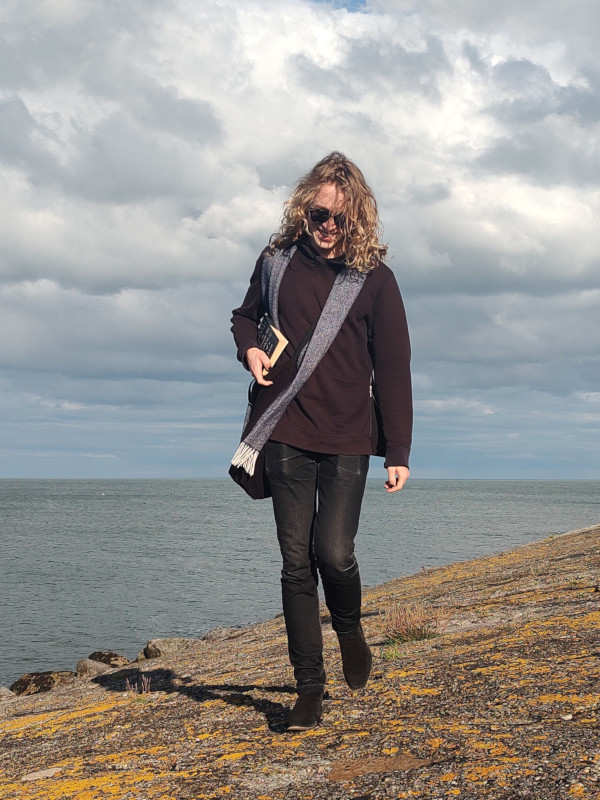 A portrait of Wiley walking by the sea with a book tucked underarm.